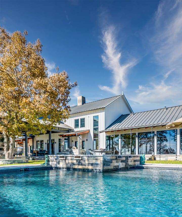 Luxury backyard pool with stone water feature in front of a modern white farmhouse with metal roof and floor-to-ceiling windows on a sunny autumn day