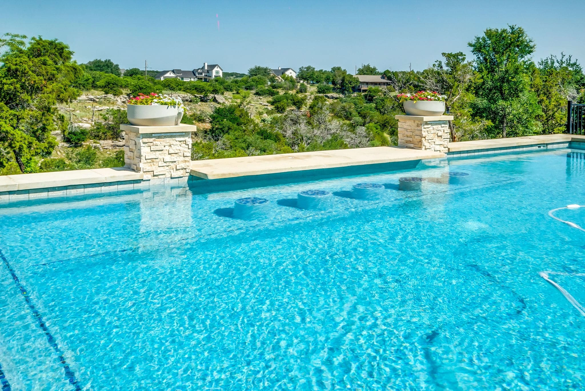 Large custom pool with in-water barstools on tanning ledge, stone columns with flower planters, and hill country views