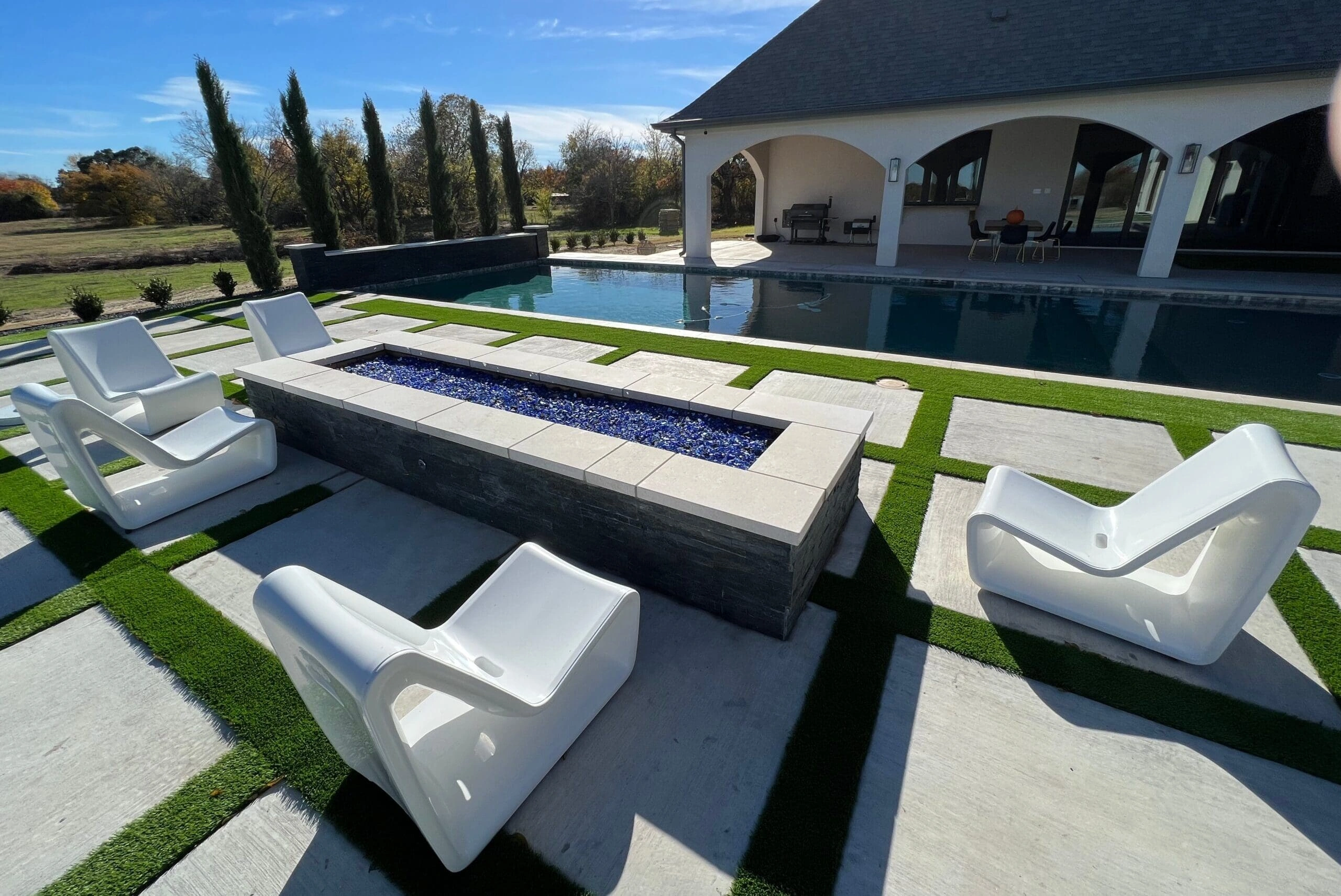 Contemporary poolside area with white lounge chairs, rectangular blue glass fire pit, and large concrete pavers in artificial turf