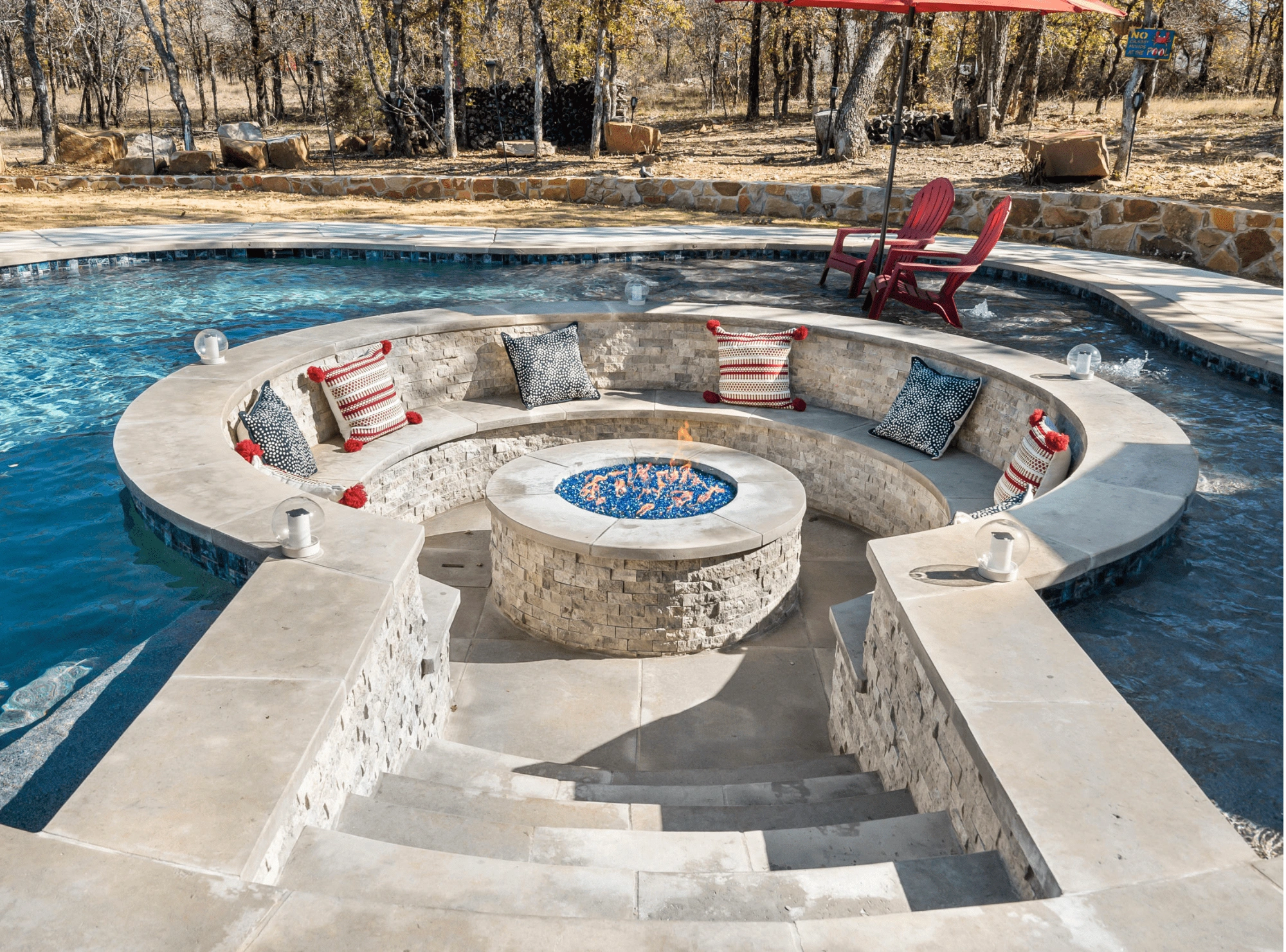 Custom pool with a built-in circular stone fire pit lounge area surrounded by blue water and decorative pillows
