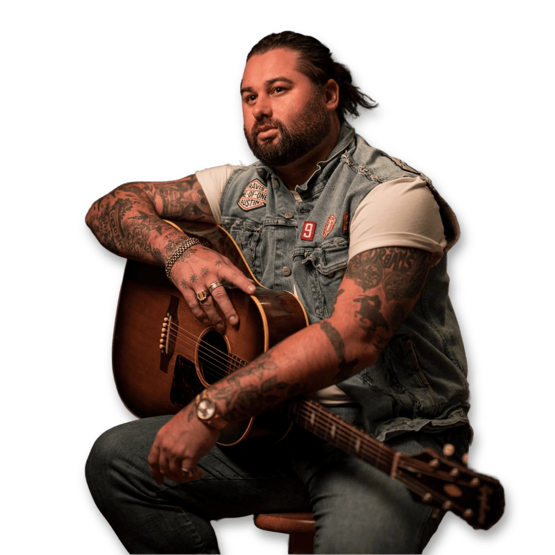Untitled-design-2025-06-05T163030.600 Tattooed musician sitting with an acoustic guitar wearing a denim vest with patches