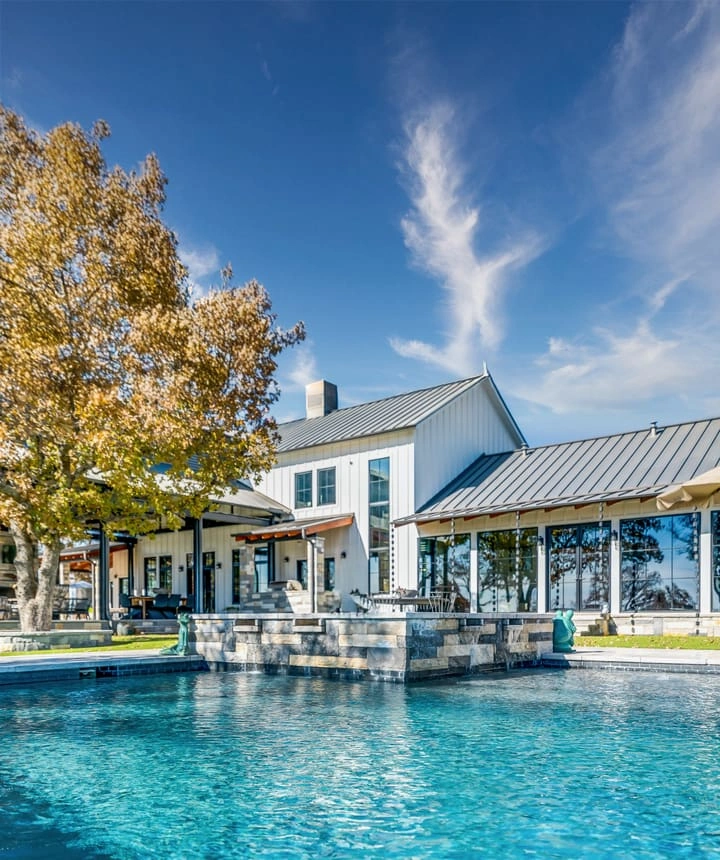 Website-House_2 Luxury backyard pool with stone water feature in front of a modern white farmhouse with metal roof and floor-to-ceiling windows on a sunny autumn day